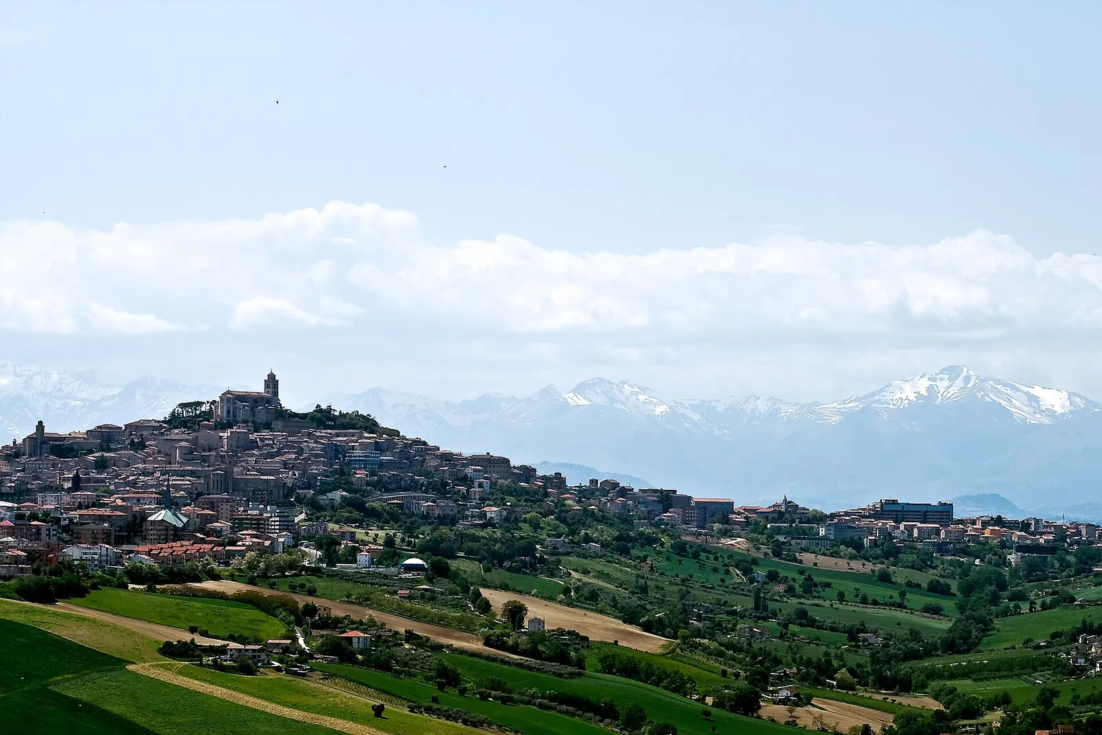 Veduta panoramica di Fermo su colle tra campagne verdi con Monti Sibillini innevati sullo sfondo