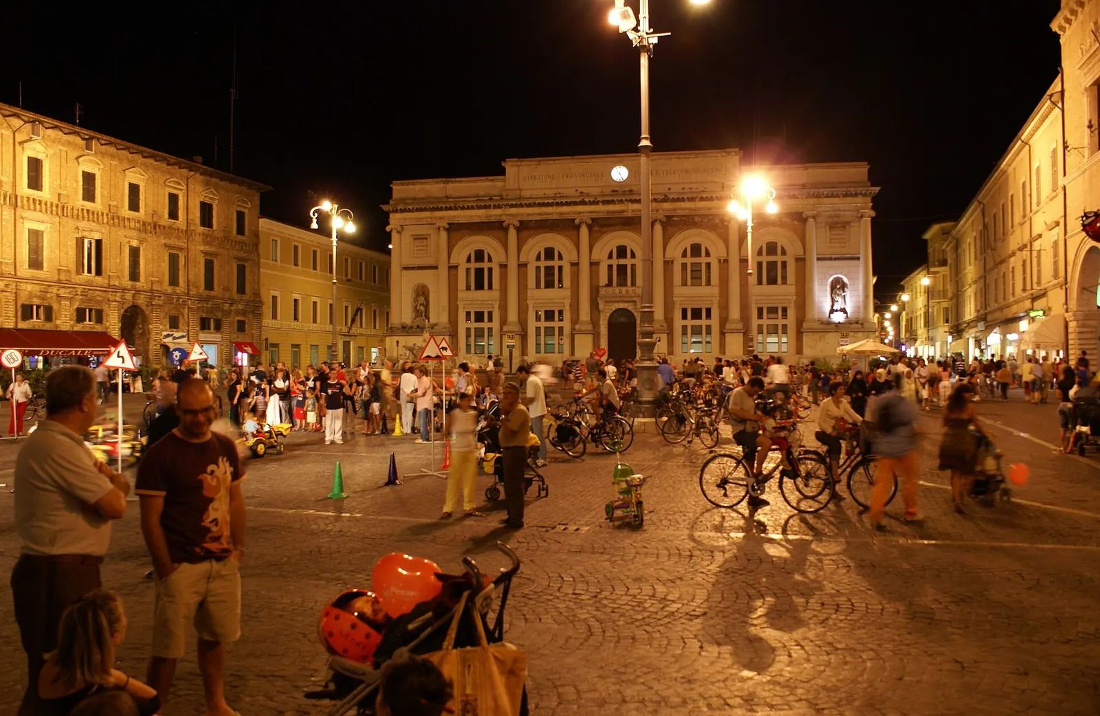 Piazza del Popolo di Pesaro animata da folla e biciclette in serata con illuminazione natalizia