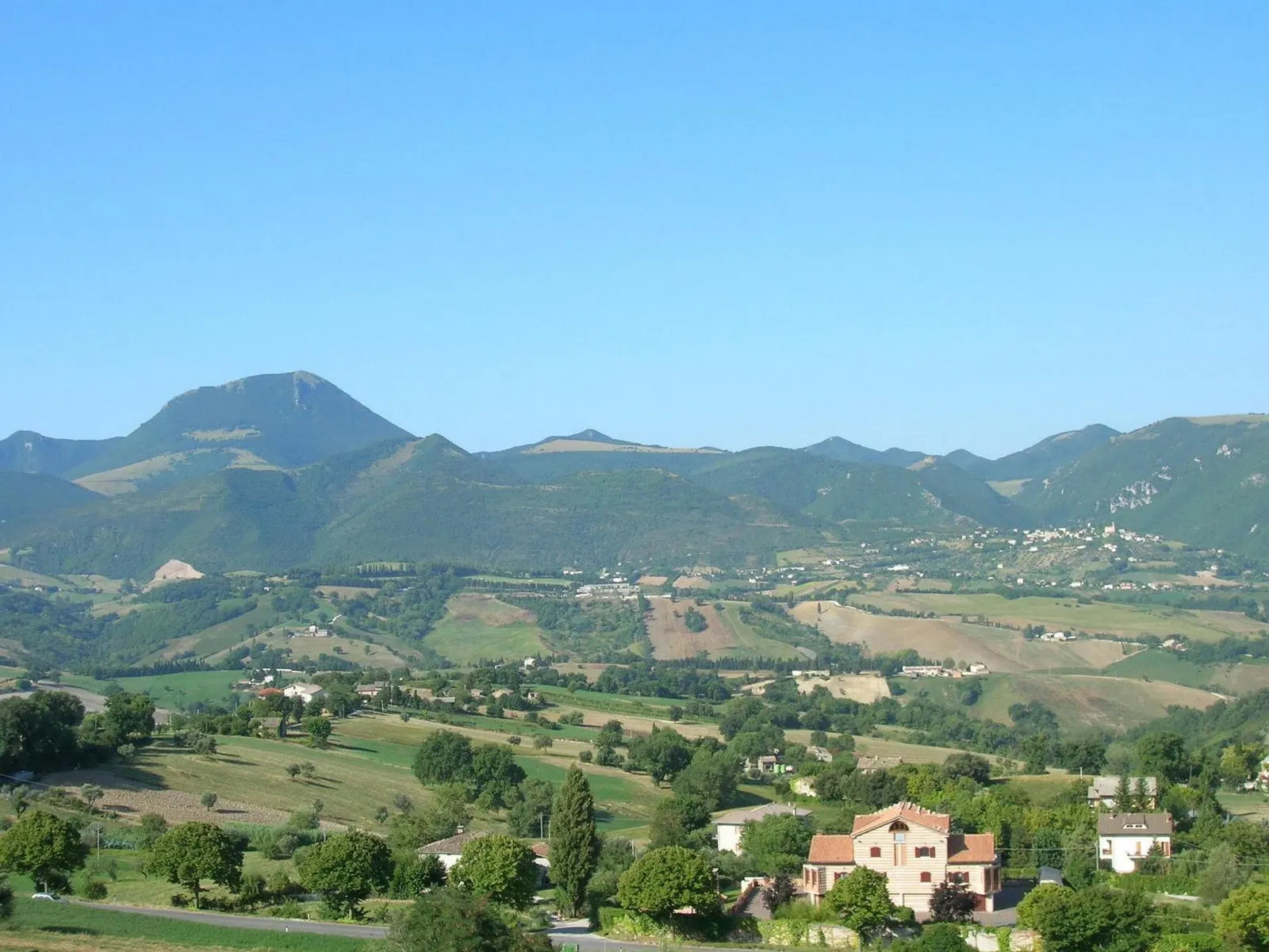 Panorama collinare di Poggio San Vicino con monte sullo sfondo e case sparse tra campi verdi in provincia di Macerata