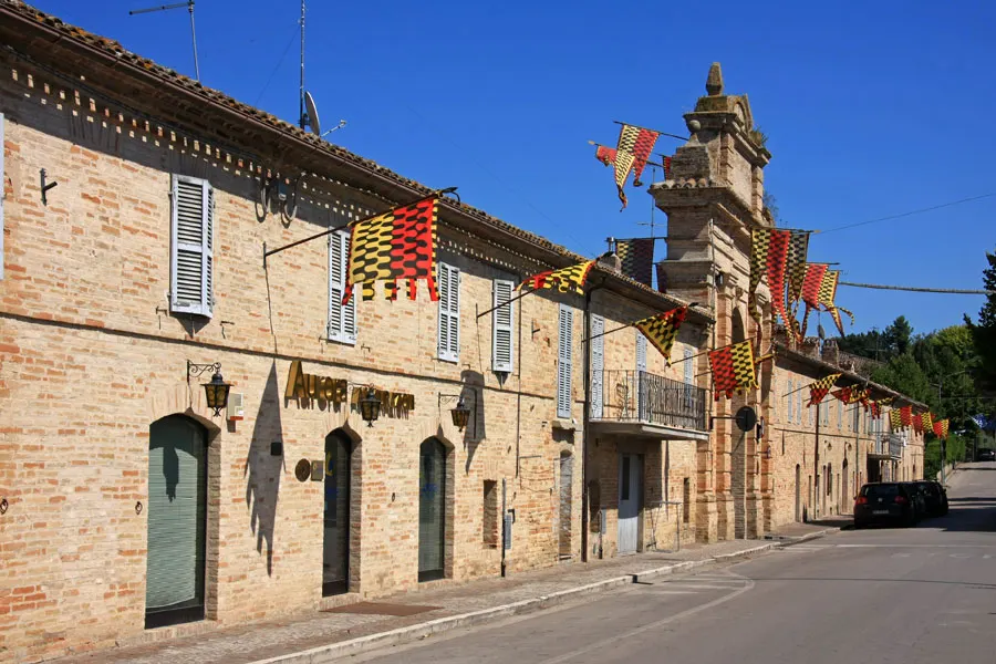 Centro storico di Servigliano con edifici in mattoni decorati da bandiere colorate e campanile sotto cielo azzurro