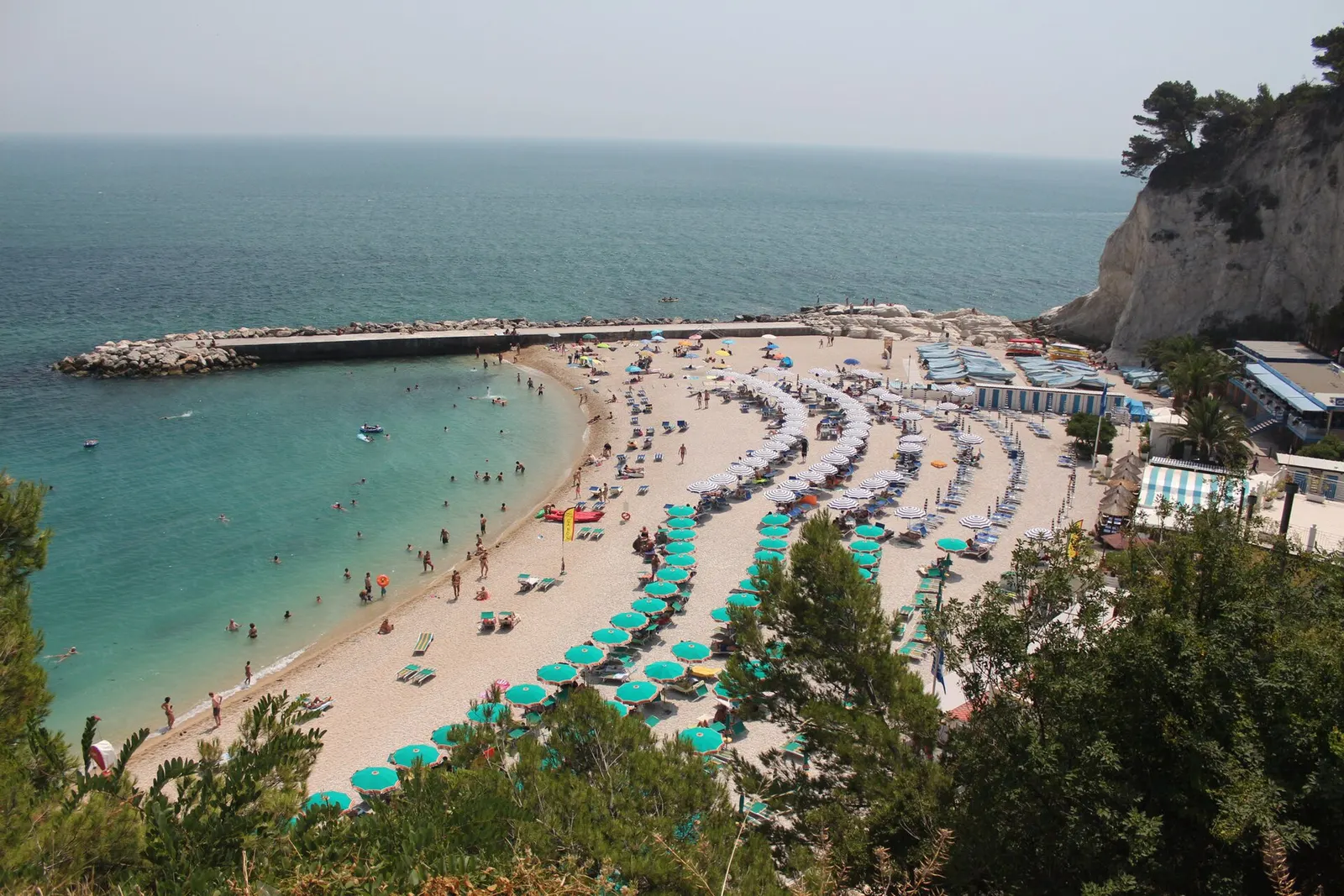 Vista panoramica della Spiaggia Urbani a Sirolo con ombrelloni turchesi e scogliera del Conero sullo sfondo